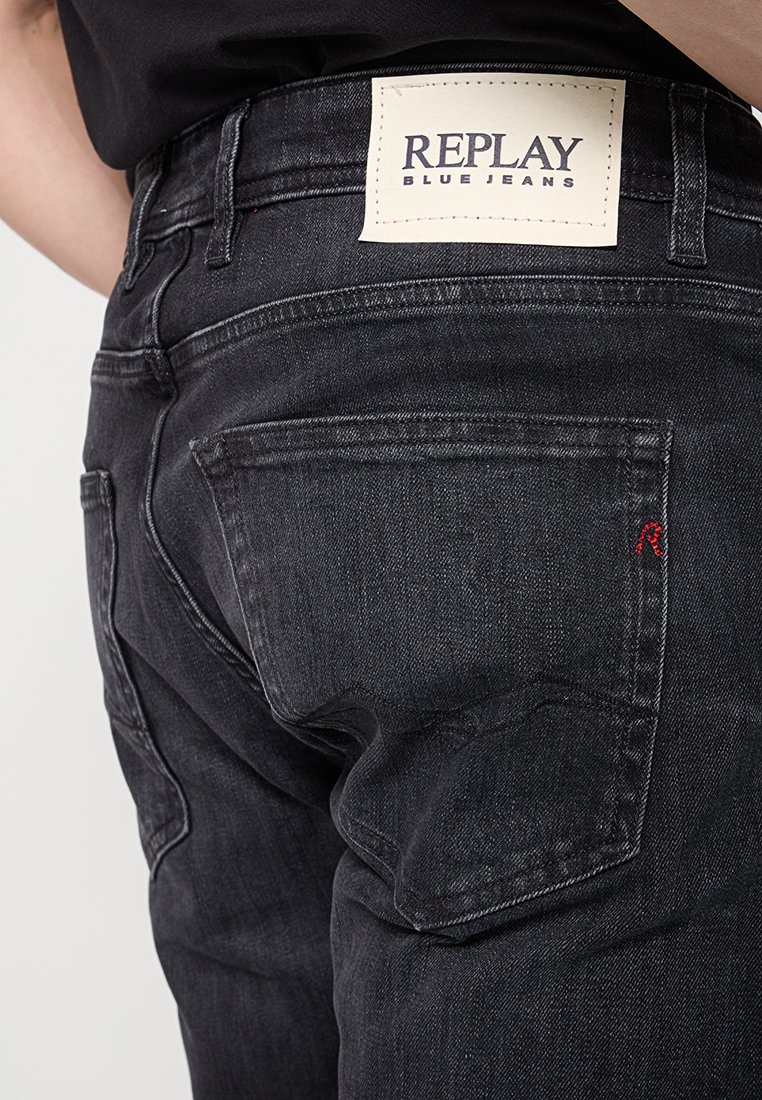 Persoon die zwarte Replay blauwe jeans draagt, met het achterzak zichtbaar en een gebrandmerkt wit leren label op de tailleband.
