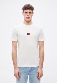 Λευκό βαμβακερό μπλουζάκι με κοντά μανίκια και στρογγυλή λαιμόκοψη· διαθέτει μικρό ορθογώνιο λογότυπο σε μαύρο και κόκκινο στο στήθος.