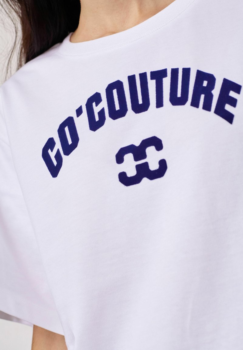 Λευκό μπλουζάκι με σκούρο μπλε κείμενο "CO'COUTURE" κυρτωμένο στο στήθος και ένα στυλιζαρισμένο λογότυπο κάτω από το κείμενο.