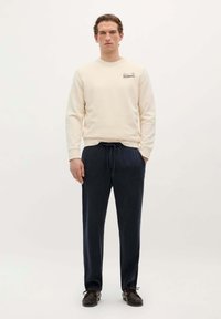 Lichtbeige sweatshirt met ronde hals en logo op de borst, gecombineerd met marineblauwe broek met trekkoord. Beide hebben een ontspannen pasvorm en een zachte textuur.