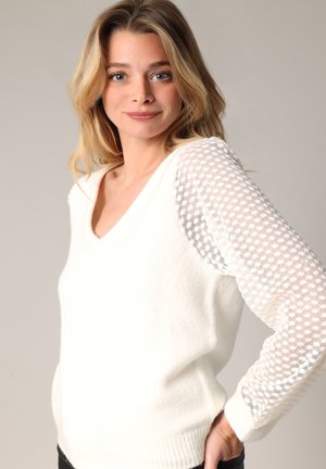 Pull blanc avec un col en V et des manches en maille transparente texturée et à motif. Matière douce, ourlet côtelé et coupe décontractée.