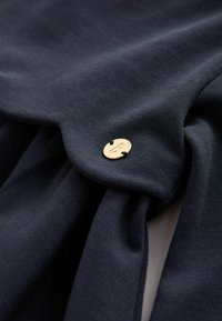 Donker marineblauwe stof met een zachte textuur, voorzien van een gouden ronde knoopdetail dat met zwarte draad is bevestigd. Geplooid ontwerp is zichtbaar.