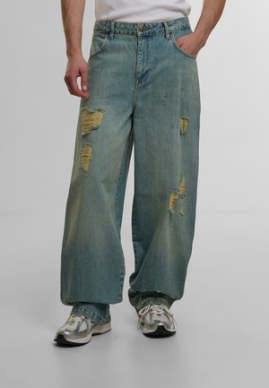 Wide-leg blue denim jeans with distressed rips and frayed edges; worn with gray and silver sneakers; five-pocket design.