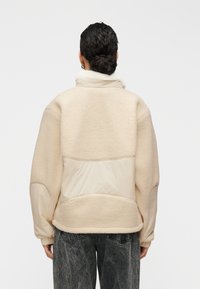 Veste en polaire beige à texture douce, dotée d'un col montant et d'un panneau arrière en tissu lisse contrastant. Coupe ample.