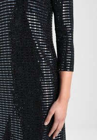 Svart klänning med metalliska paljetter i olika storlekar placerade i horisontella rader, trekvartsärmar och en figurnära silhuett.