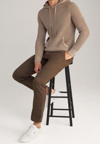 Mann in beiger Strickkleidung, braunen Hosen und weißen Sneakers, der auf einem hohen schwarzen Hocker vor hellem Hintergrund sitzt.