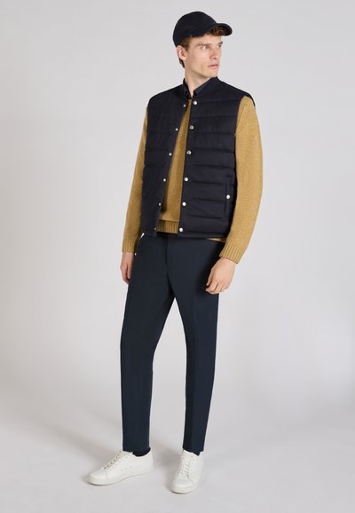 Gilet rembourré bleu marine sur un pull beige, associé à un pantalon sombre et des baskets blanches. Le gilet est doté de boutons-pression et d'un col.