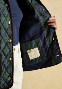 Jachetă matlasată de culoare verde închis, cu detalii maro; căptușită cu țesătură bleumarin cu motive tartan. Include o etichetă cu instrucțiuni de îngrijire și numele brandului.