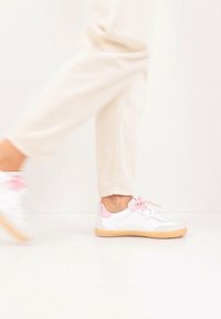 Λευκά αθλητικά παπούτσια με ροζ λεπτομέρειες και κορδόνια, με λεία, υφή επιφάνεια και σόλα από καουτσούκ. Τα παπούτσια φοριούνται με ανοιχτόχρωμο παντελόνι.