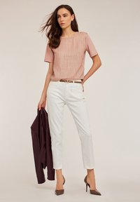 Top roz deschis cu un model texturat, pantaloni albaștri slim-fit și curea maro. Modelul ține o geacă burgundă, purtând tocuri maro ascuțite.