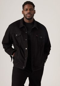 Mustast teksajope klassikalise disainiga, millel on kaks rinnataskut, hõbedased nupud ja lõdvestunud lõige. Kantud musta t-särgi peal.