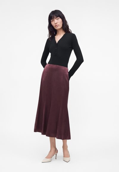 Femme portant un haut noir à manches longues, une jupe midi violet foncé et des chaussures blanches à talons, debout les mains derrière le dos.
