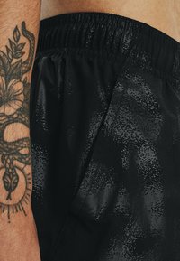 Arm met slang- en bloemtattoo naast zwarte shorts met textuur, glanzend patroon en elastische tailleband.