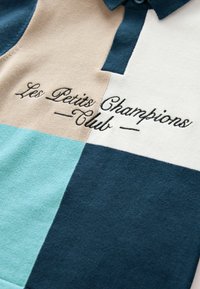 Pikétröja med en färgblockdesign i beige, teal, vit och mörkblå med broderad text "Les Petits Champions Club."