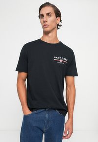 Ung mand med kort, mørkt hår iført en sort GANT 1949 T-shirt og blå jeans, stående med den ene hånd i lommen mod en hvid baggrund.