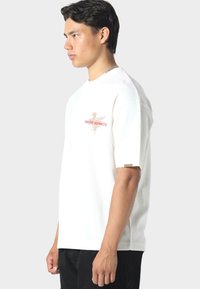 Camiseta blanca de manga corta con un diseño gráfico rojo en el pecho que presenta el texto "PAS DE MONACO". Corte simple y relajado, textura suave.