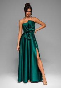Abito in raso verde smeraldo con design senza spalline, dettaglio in vita annodato e uno spacco alto fino alla coscia; presenta una texture morbida e fluente.