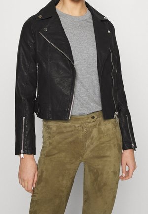 Persona con chaqueta de cuero negra de motociclista, camiseta gris de cuello redondo y pantalones de ante verde oliva, de pie contra un fondo liso.