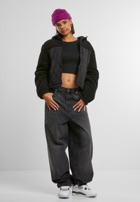 Schwarze gesteppte Jacke aus flauschigem Material, schwarzes Crop-Top, dunkle baggy Jeans und weiße Sneakers mit grünen Akzenten, dazu eine lila Mütze.