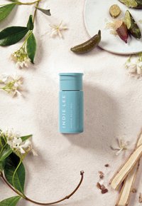 Pojemnik z niebieską lotioną oznaczoną "INDIE LEE GENTLE DAILY PEEL", umieszczony na jasnym piasku, otoczony białymi kwiatami i zielonymi liśćmi.