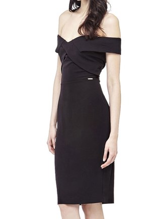 Kvinde iført en tætsiddende, sort off-shoulder kjole med indpakket overdel og knælængde, stående med armene afslappet ned langs siderne.