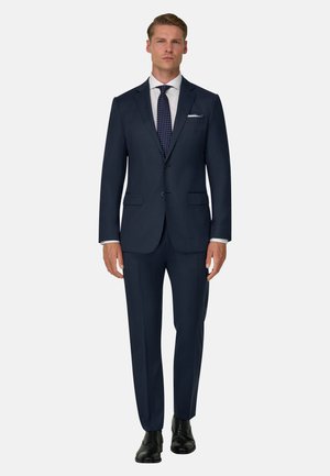 Hombre de pie, erguido, vestido con un traje azul oscuro de dos botones, camisa blanca, corbata estampada, pañuelo de bolsillo y zapatos de vestir negros sobre un fondo liso.