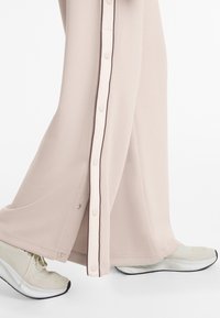 Hellebeige, weit geschnittene Hosen aus weichem Stoff mit einer weißen Knopfleiste und dunkler Naht. Getragen mit hellen sportlichen Schuhen.