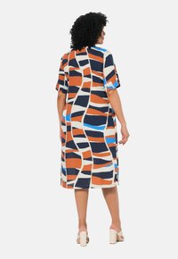 Monivärinen mekko, jossa on abstrakti kuvio oranssissa, laivastonsinisessä ja sinisessä. Lyhyet hihat frilliyksityiskohdilla ja suora leikkaus.