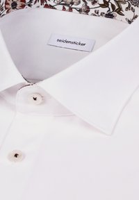 Chemise blanche à col structuré, fermeture à un bouton, avec une doublure intérieure du col à motif floral. Étiquette de marque visible.