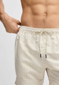 Beige gemusterte Badeshorts mit einem elastischen Bund, verstellbarem Kordelzug und schwarzen Metallspitzen am Kordelzug.
