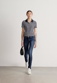 Pruhované námornícke modré a biele polo tričko s golierom, doplnené priliehavými tmavými džínsami a bielymi teniskami. Drží malú čiernu kabelku.