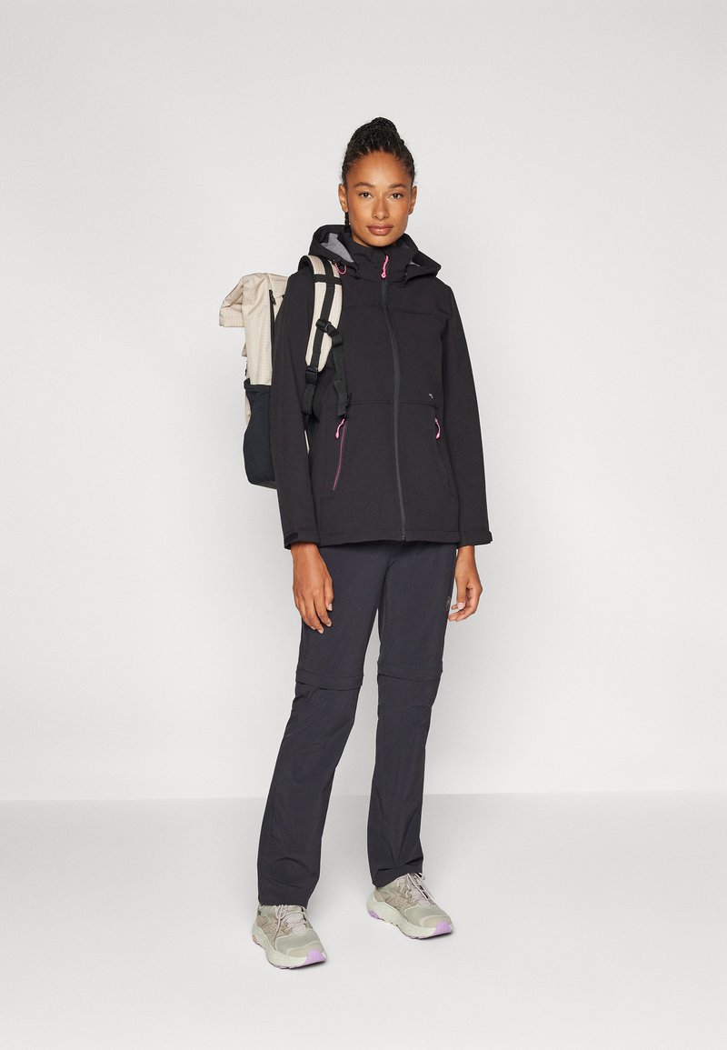 Schwarze Fleecejacke mit Frontreißverschluss und Kapuze, kombiniert mit dunkelgrauen Hosen. Die Frau trägt einen beige-schwarzen Rucksack und graue Sneaker.