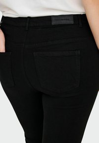 Crne traperice od denima s uskim krojem, sadrže dva stražnja džepa i crnu oznaku na struku, ističući glatku teksturu.