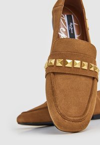 Mocassins en daim marron à semelle plate, ornés d'une bande décorative de clous pyramides dorés sur le dessus. Surface texturée avec des détails cousus.
