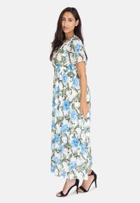 Blommig maxiklänning med vit bas, blå blommor och gröna blad. Korta ärmar, åtsittande midja och flödande silhuett. Svarta klackskor.