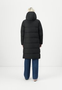Svart pufferjakke med hette, med kviltet design og lang lengde, kombinert med vide denimjeans og sandaler.