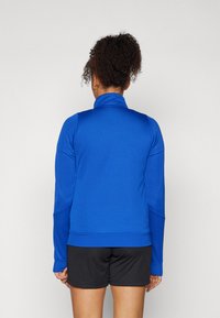 Blue athletic jacket in smooth, stretchy material with a high collar and long sleeves, paired with black shorts. Back view shows seamless design.