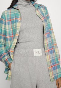 Rutig skjorta med knappar i blått, grönt och gult, lagd över en grå turtle neck. Grå sweatpants har en "POLO"-logotyp.