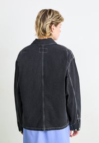 Črna denim jakna s sproščenim krojem, ki ima kontrastno belo šivanje, zapenjalne manšete s gumbnico in ovratnik.