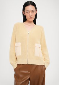 Cardigan giallo a costine con scollo a V e zip argentata, dotato di due tasche a toppa color crema. Vestibilità leggermente oversize.