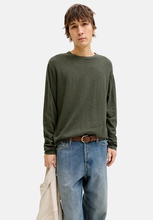 Jonge persoon met kort bruin haar, gekleed in een olijfgroen shirt met lange mouwen en een blauwe spijkerbroek, met een beige jas in één hand.