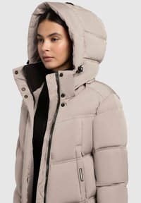 Bežs pildīts apģērbs ar augstu apkakli un lielu kapuci, ar rāvējslēdzēju un pogu aizdari, vairākiem kabatām un quilted faktūru.