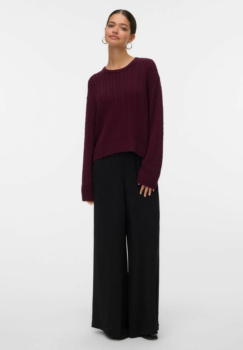 Maglione lavorato a maglia borgogna con una trama a costine, caratterizzato da un collo rotondo e spalle scese, abbinato a pantaloni neri larghi a gamba larga.