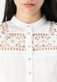 Camicia bianca con colletto alla mandarino, caratterizzata da delicate rifiniture in pizzo e bottoni sul davanti. Tessuto leggero e strutturato con motivi discreti.