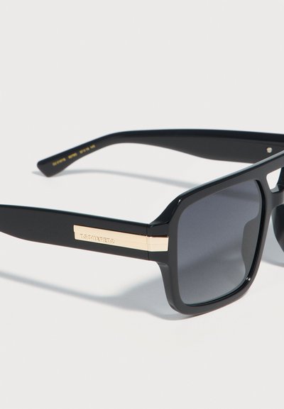 Μαύρα γυαλιά ηλίου με τετράγωνο σκελετό, σκούρους φακούς και χρυσές λεπτομέρειες στα πλαϊνά μέρη, με χαραγμένο το όνομα της μάρκας.