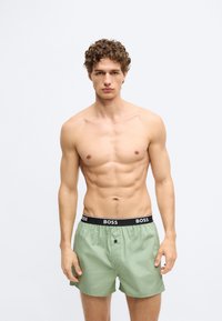 Zelené plavkové šortky s čiernym pásom nesúcim názov značky "BOSS". Ľahká látka s hladkým povrchom a detailom s predným gombíkom.