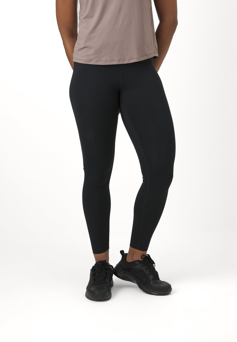 Leggings neri con una texture liscia, caratterizzati da un design aderente e cuciture a contrasto. Indossati con un top marrone chiaro e scarpe sportive nere.