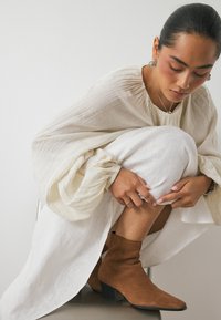 Bottes de cheville en daim marron avec un bout pointu et un talon empilé, associées à une robe blanche fluide et un haut crème texturé. La lumière naturelle met en valeur les détails.
