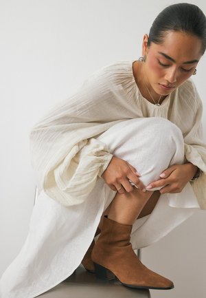Bruna mockakängor med spetsig tå och staplad klack, kombinerade med en flödande vit klänning och en texturerad krämfärgad överdel. Naturligt ljus framhäver detaljer.