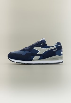 Tengerészkék és szürke sportcipő fűzővel, márkás oldalsávval és fehér talppal, semleges háttéren, külső oldalról nézve.